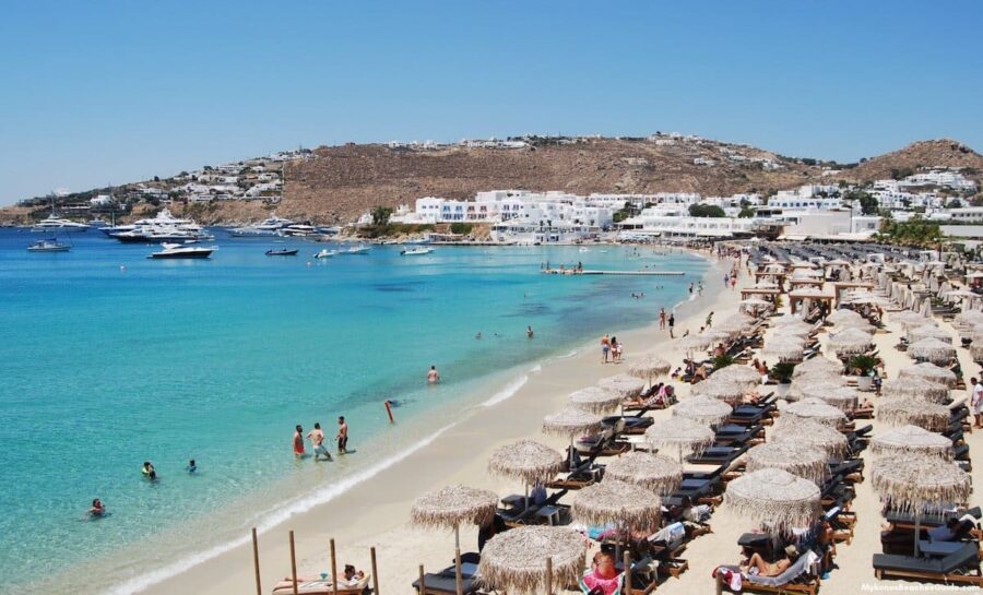 Vibrant Greek beach scene with sun loungers, turquoise waters, and hillside whitewashed buildings.