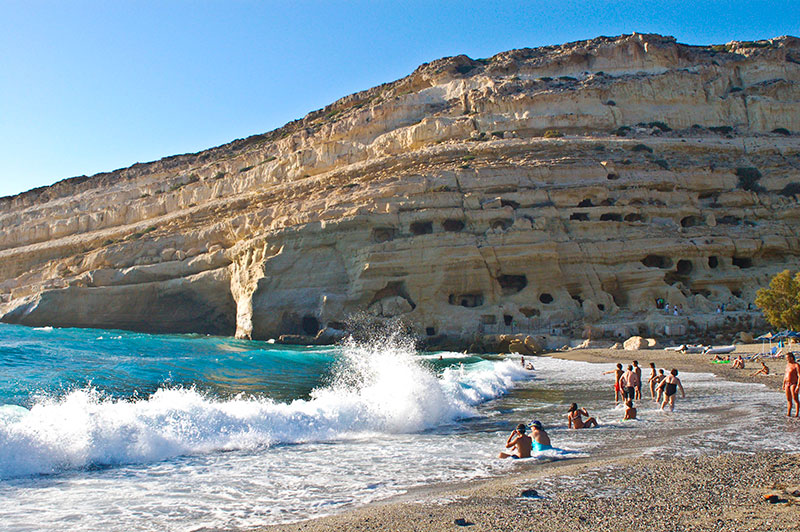 Serene beach scene with cliffs, azure sky, waves, sunbathers, and turquoise water at Matala Beach.
