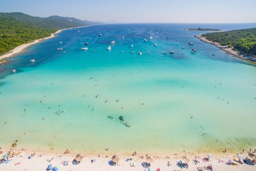 Tranquil Sakarun Beach: turquoise waters, sunbathers, colorful umbrellas, boats, and lush hills.