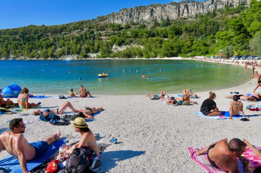 Serene beach scene in Split, Croatia with sunbathers, azure waters, and lush greenery.