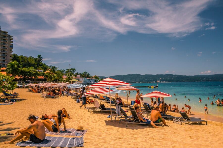 Sunny beach scene with sunbathers, striped umbrellas, turquoise ocean, and lush tropical surroundings.