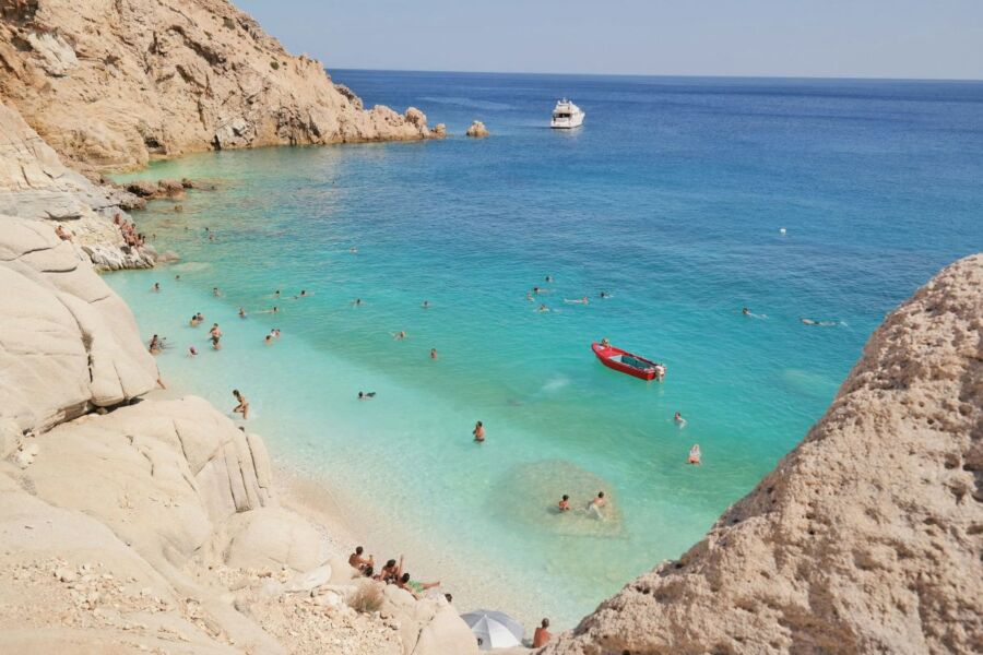 Sunny beach scene with turquoise waters, rocks, boats, and people enjoying the coastline.