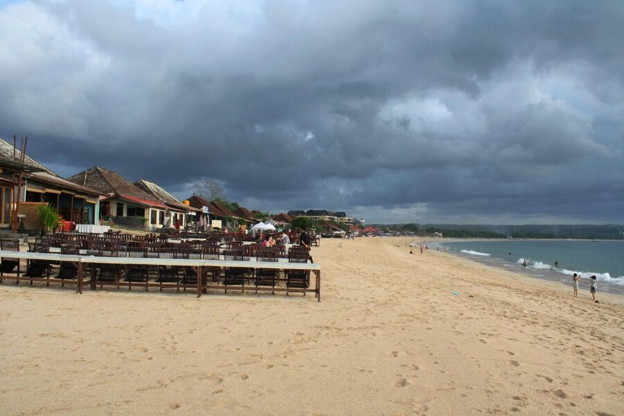 Serene Jimbaran beach scene with dining area, ocean waves, and a vibrant sunset sky.