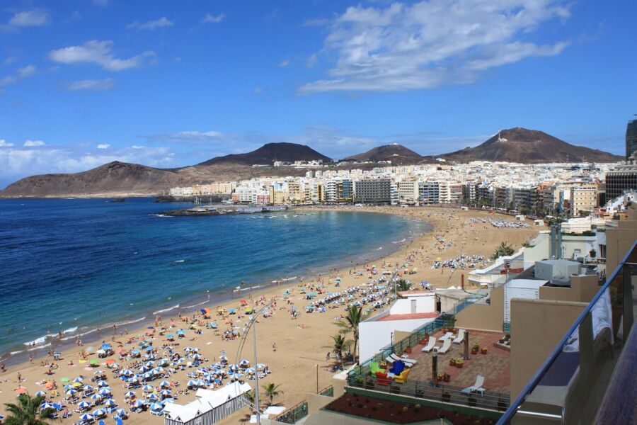 Vibrant Las Palmas beach scene with sunbathers, ocean, pastel buildings, and scenic mountains.