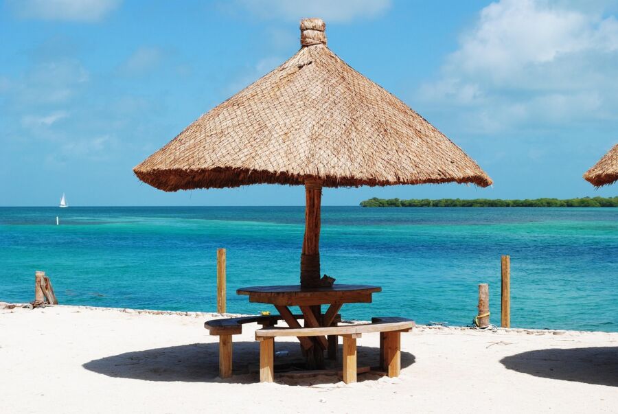 Serene beach scene with rustic table, thatched umbrella, turquoise ocean, and sailboat.