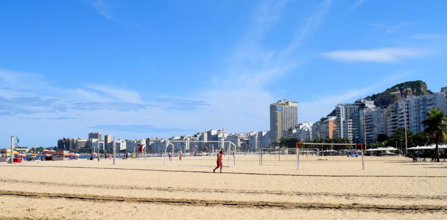 Bustling beach with sand, volleyball, umbrellas, people, modern buildings, and mountain backdrop.