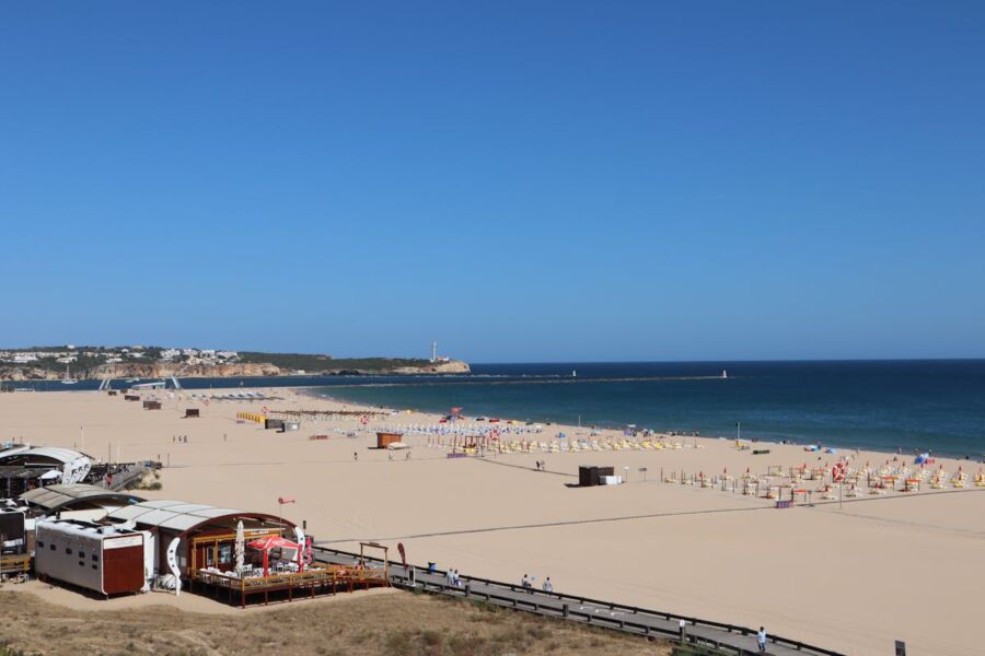 Beautiful Portimao beach with golden sands, colorful umbrellas, clear sky, and distant lighthouse.
