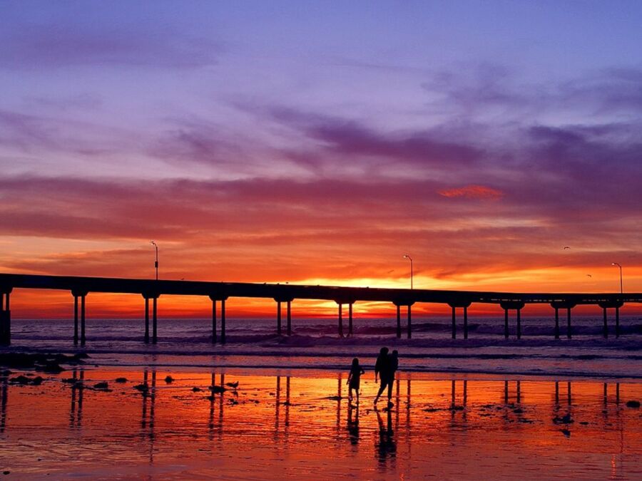 Sunset beach scene with pier, silhouetted figures, and vibrant sky reflections on the water.
