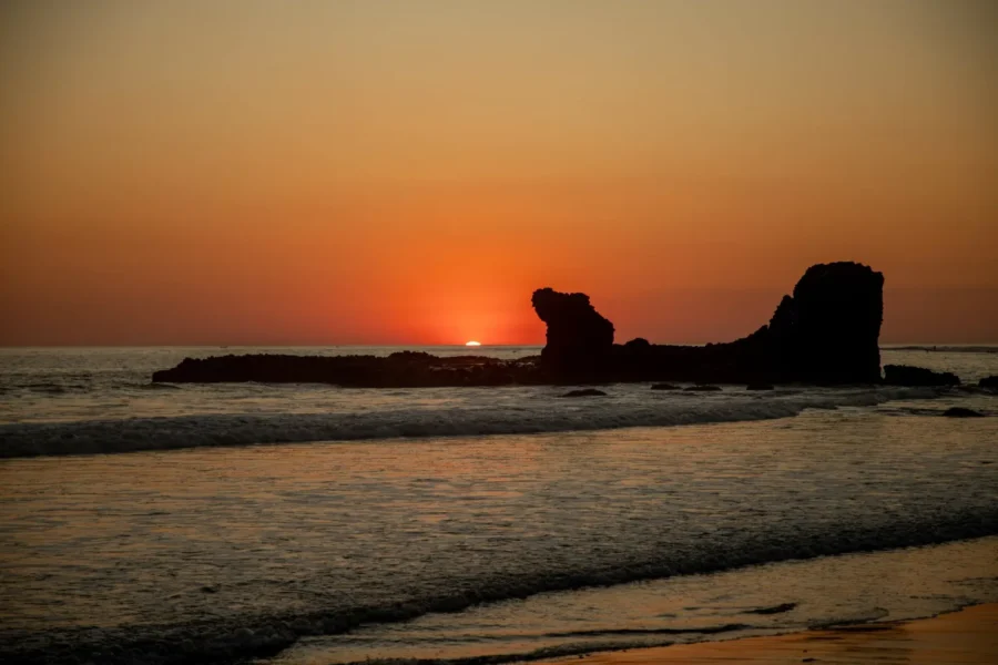 Beach sunset with colorful sky, serene waves, and silhouetted rock formations.