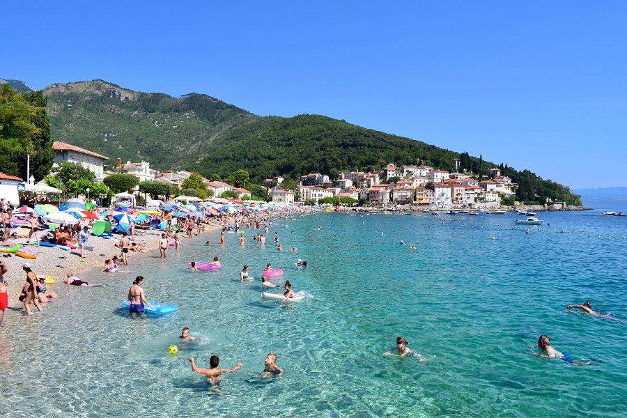Vibrant beach scene with umbrellas, swimmers, palm trees, and picturesque Mediterranean town.