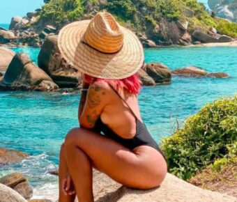 Woman in straw hat with pink hair sits on coastal rocks overlooking turquoise ocean.