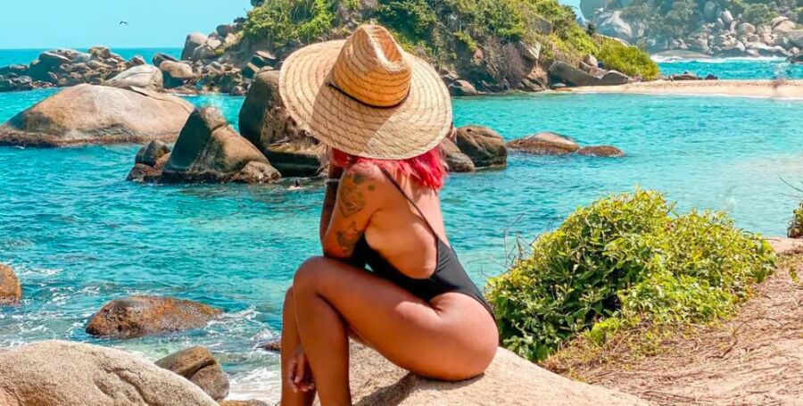 Woman in straw hat with pink hair sits on coastal rocks overlooking turquoise ocean.