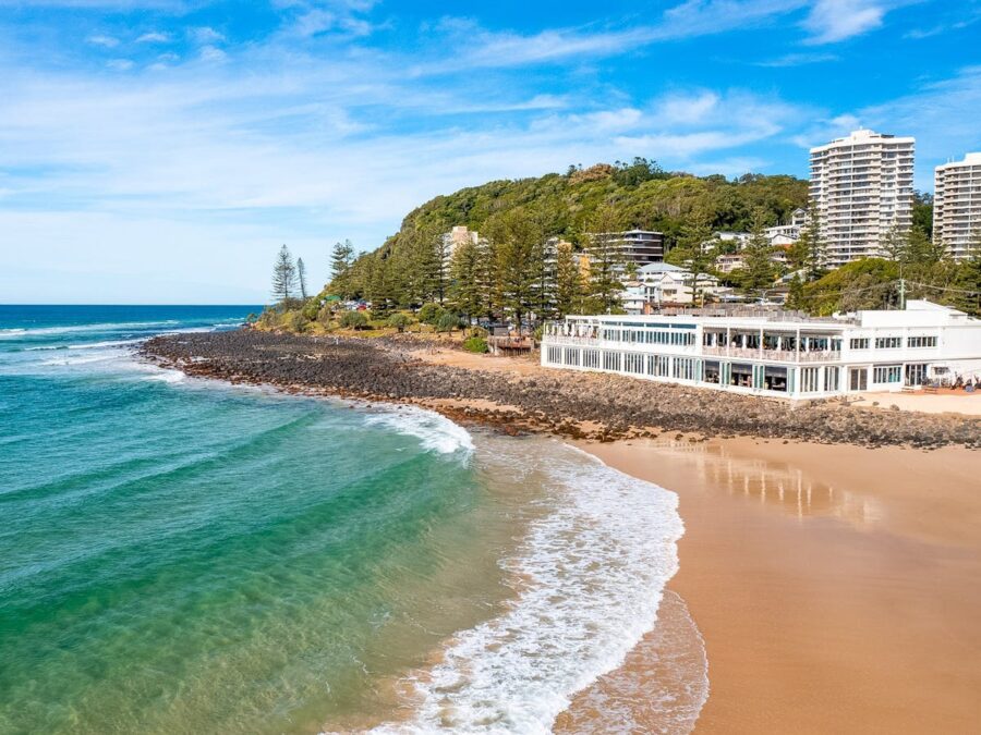 Burleigh Heads beachfront resort with turquoise ocean waves, sandy beach, coastal apartments and lush green hillside.