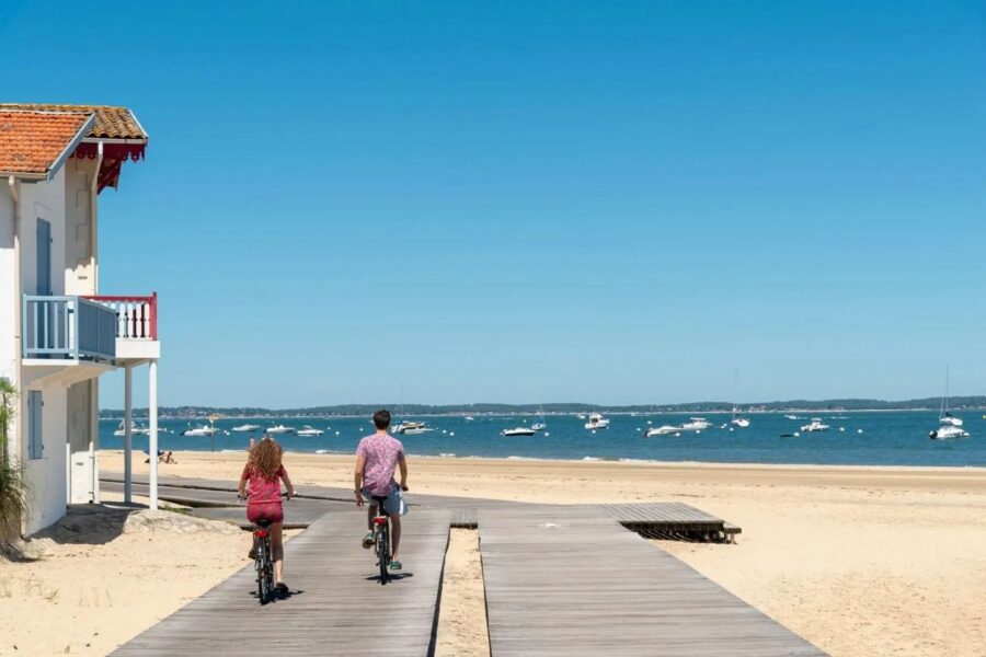 A vibrant beach scene at Arcachon Bay unfolds under a clear sky, with a golden shore, white beach house, cycling duo on a boardwalk, and tranquil boats on the sea. The setting embodies relaxation and joy, perfect for a sunny day by the sea.