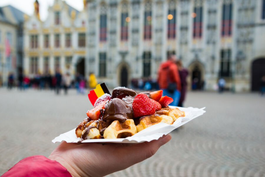 Hand holding a Belgium waffle topped with chocolate sauce and strawberries, with Bruges cityscape in the background.