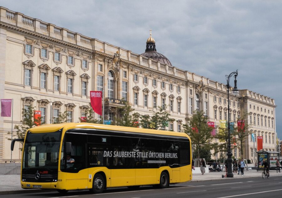 Yellow city bus in front of historic building with European architecture, city life bustling around.