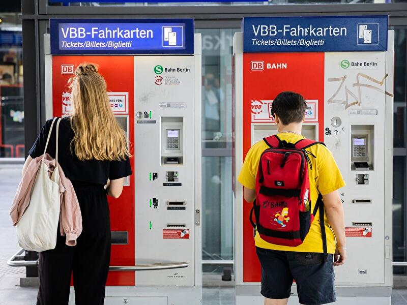 Two travelers using Berlin transit ticket machines at a busy multicultural station.