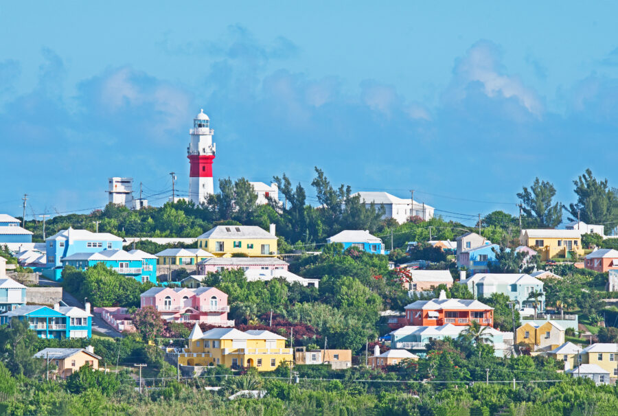 Colorful pastel homes line the hillside in Bermuda, topped by a red and white lighthouse against a clear blue sky.
