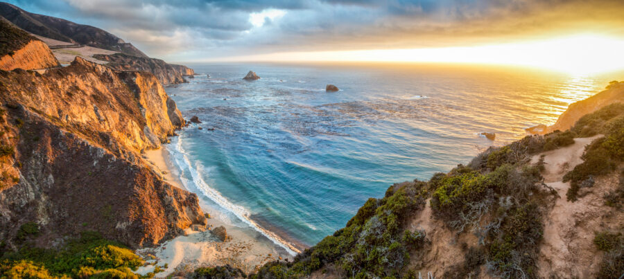 Sunset panorama of Big Sur coastline, highlighting dramatic cliffs and the ocean's shimmering surface in California.
