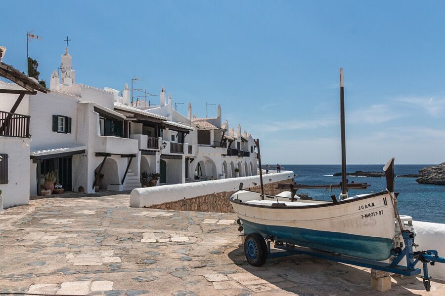 Boat parked beside a road in Binibeca, Menorca, surrounded by greenery and coastal scenery.