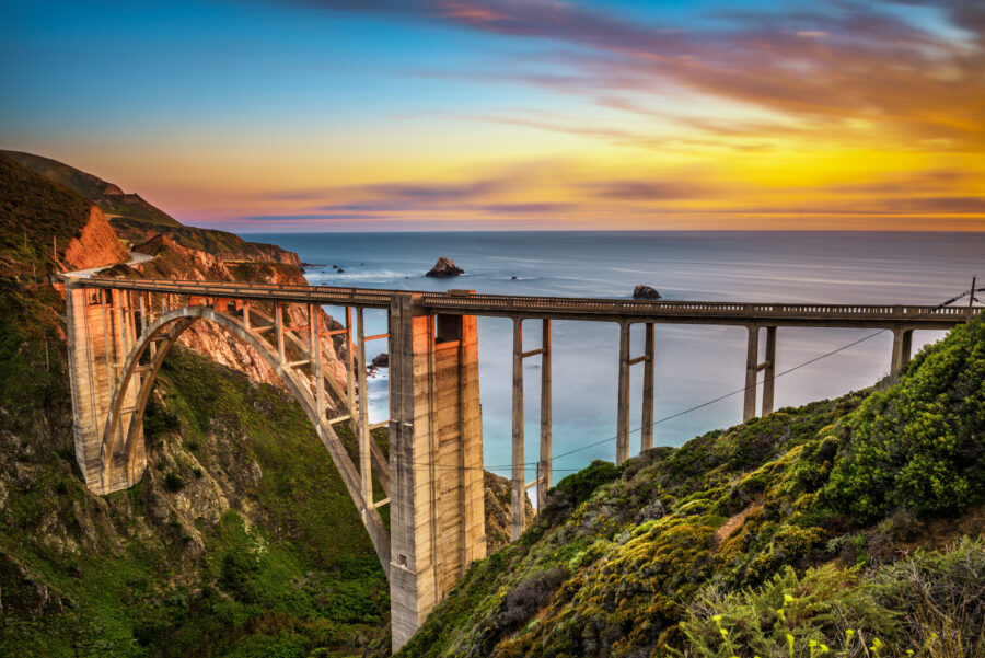 Bixby Bridge and the Pacific Coast Highway are bathed in sunset colors, showcasing the beauty of the California coastline.


