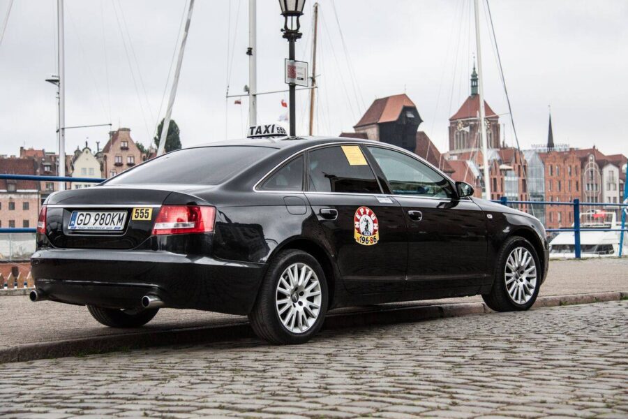 Black sedan taxi parked on Gdansk waterfront promenade with harbor and historic buildings.