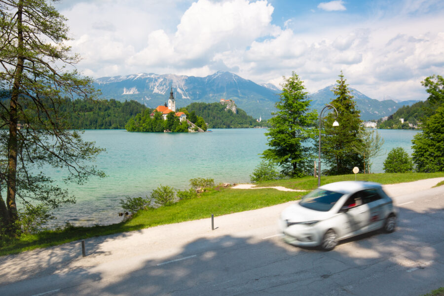 White car drives along a scenic road by a beautiful blue lake in Slovenia, surrounded by lush greenery