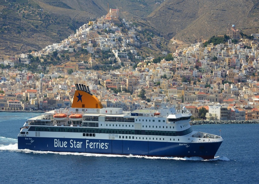 Blue Star ferry sailing near picturesque Greek island town with vibrant architecture and lush greenery.