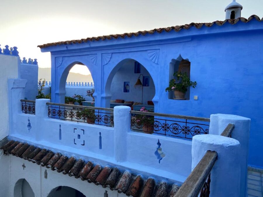 Blue Moroccan terrace balcony at sunset with arches, potted plants, mountains and vibrant painted walls.