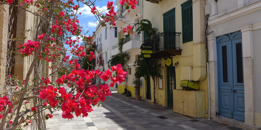 Nafplio cobblestone alley with bougainvillea, pastel buildings, balconies, and a sunny cafe entrance.