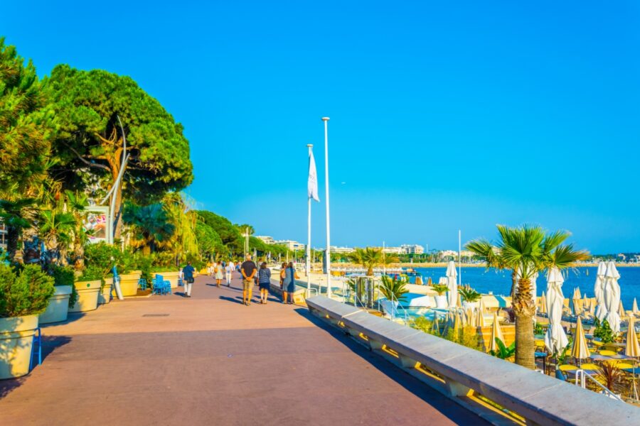 People leisurely walking along Boulevard de la Croisette in Cannes, France, with palm trees and shops in the background