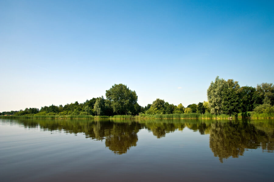 Tranquil Bovenwijde Giethoorn lake, framed by lush trees and a bright sky, reflecting nature's calm beauty.
