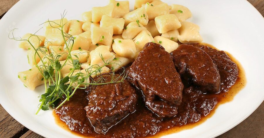 Braised beef stew with potato gnocchi, garnished with green onions and microgreens on a wooden table.