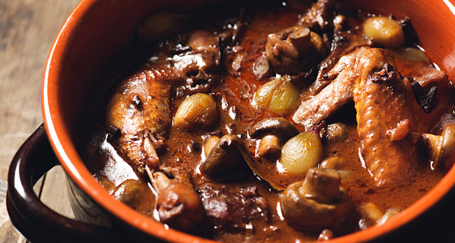 Braised chicken coq au vin with mushrooms and pearl onions in rustic earthenware pot.