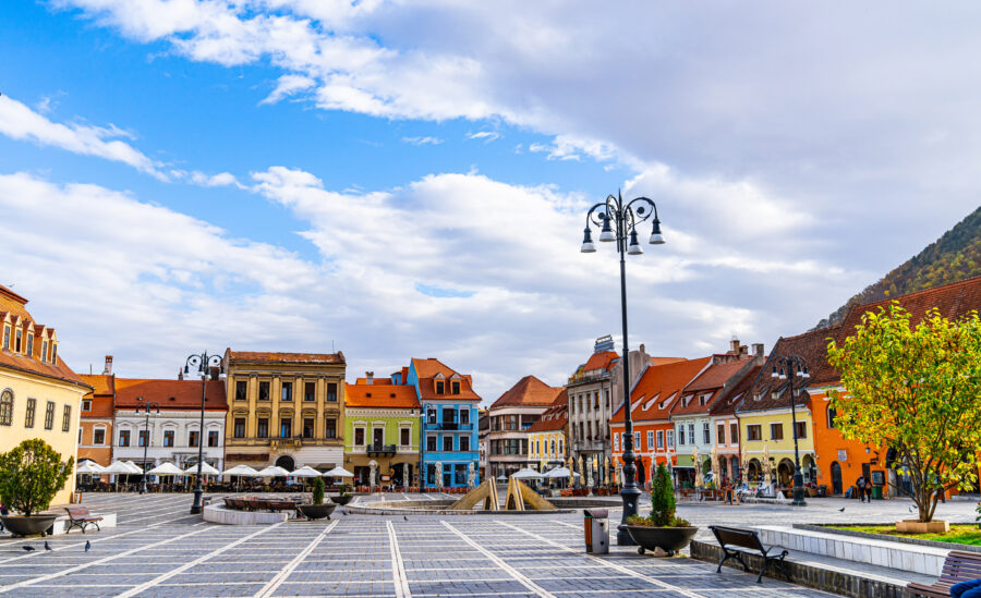 Bright square in Brașov, Romania, filled with colorful architecture, outdoor dining, and a clear sky, perfect for visitors.