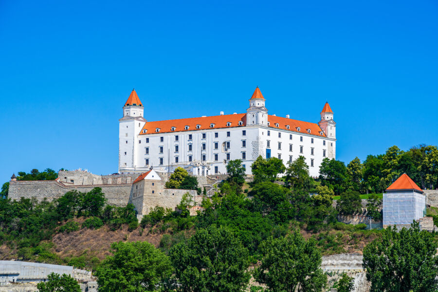 Bratislava Castle, a medieval fortress, stands prominently on a hill, surrounded by lush greenery and a vibrant skyline