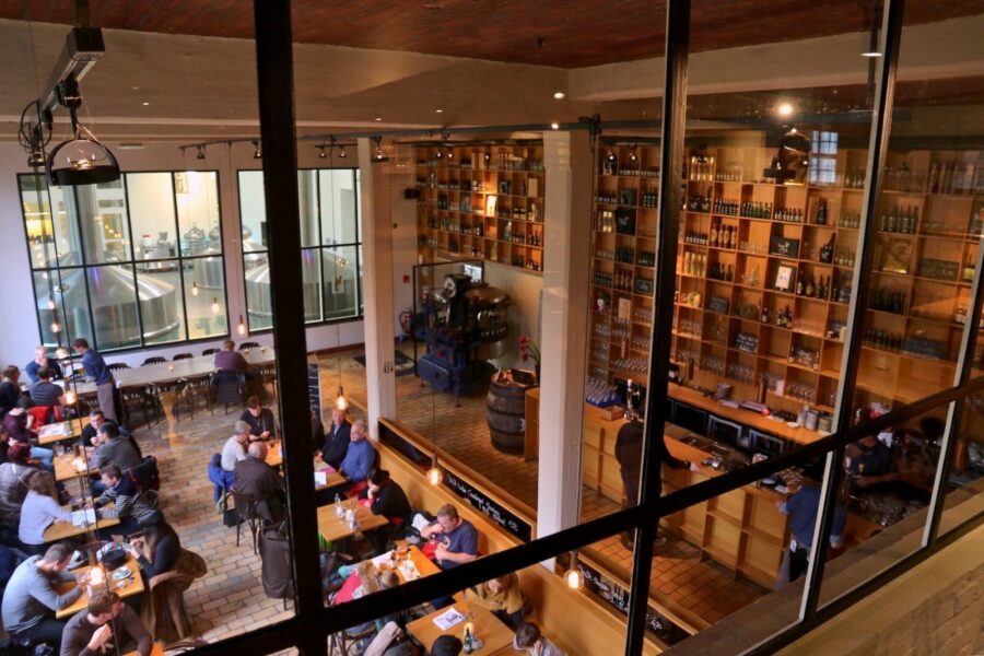 Taproom interior with communal wooden tables, brewing tanks, bar shelving, warm industrial lighting.