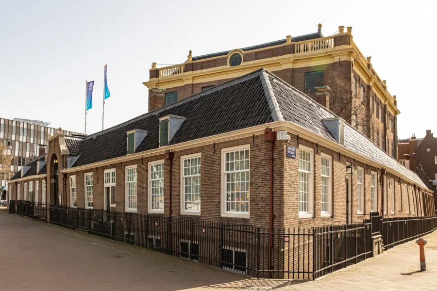 Historic brick building complex with dormer roof, street signs, metal fence and modern city backdrop.