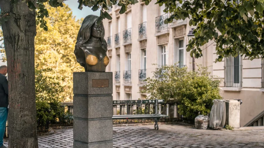 Bronze bust of Dalida on granite pedestal in shady urban square.
