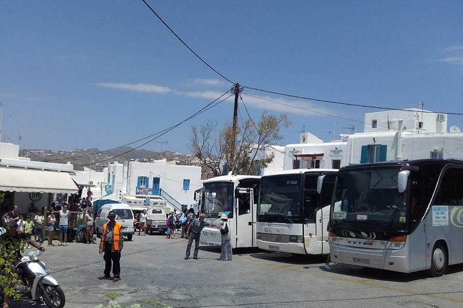 Bustling Greek island square with white tourist buses, vibrant buildings, and lively Mediterranean ambiance.