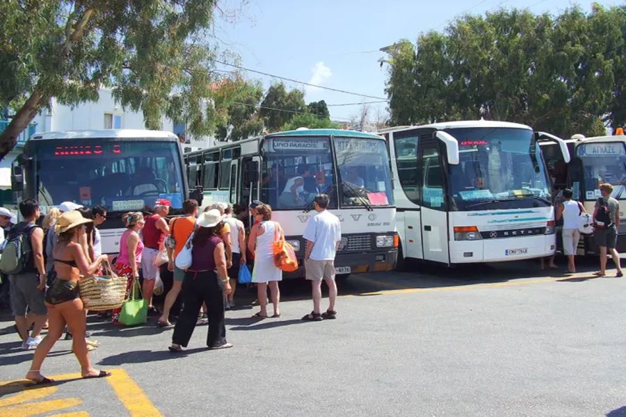 Bustling Mykonos bus station: travelers in summer attire await buses under sunny skies.