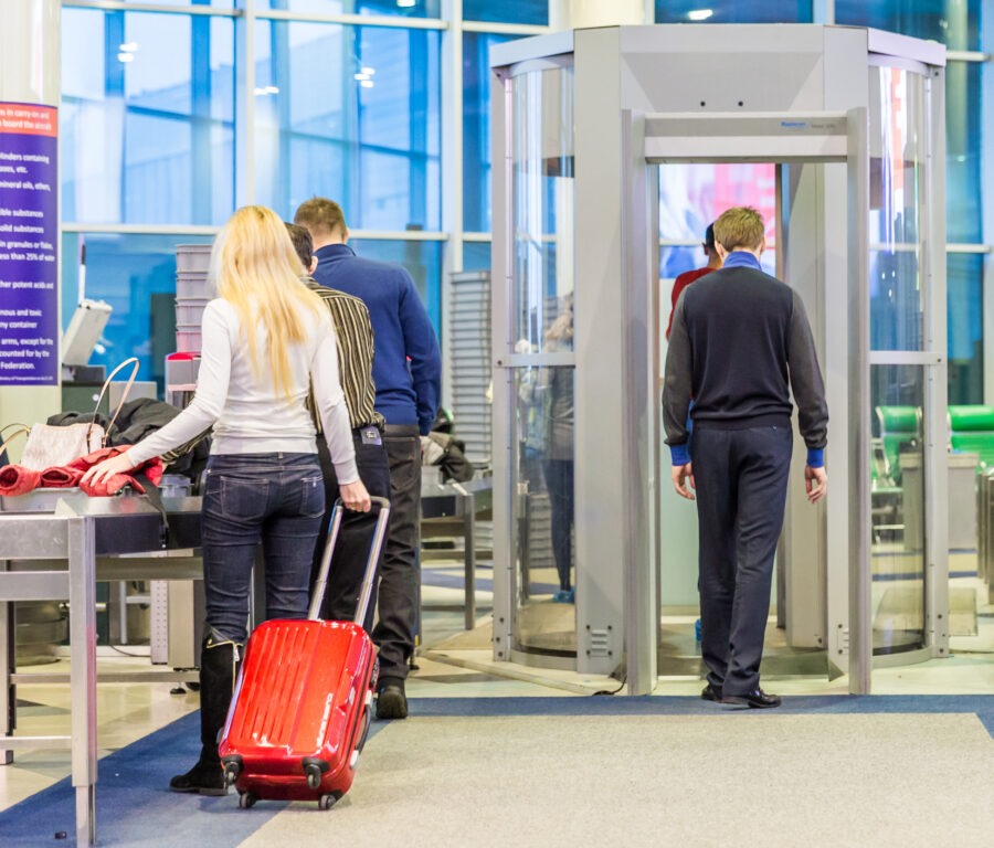 Crowded airport security area with people queuing, security personnel checking luggage and assisting travelers.