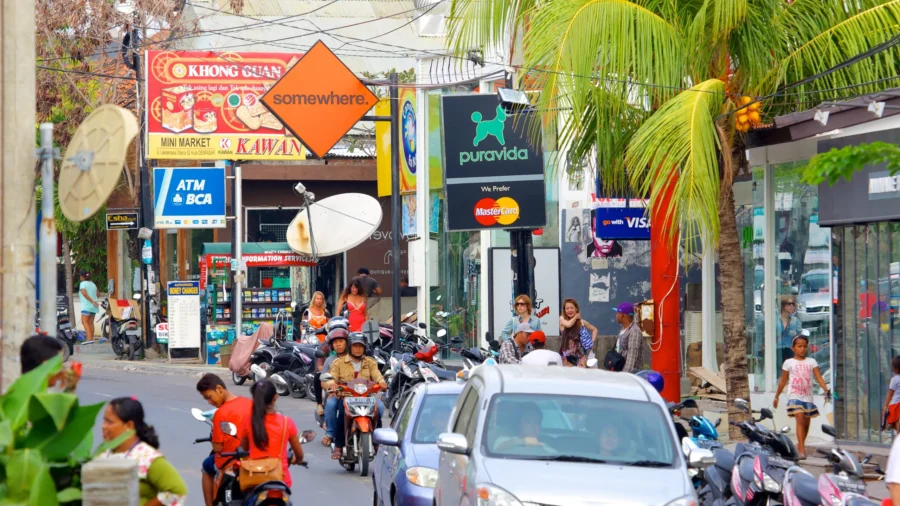 Busy Seminyak Bali street with motorbikes, cars, shops, palm trees, tourists, and local traffic.