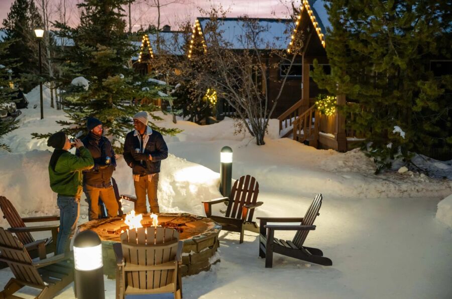 Four people gather around a fire pit in the snow, with cozy cabins visible in the background at Yellowstone.