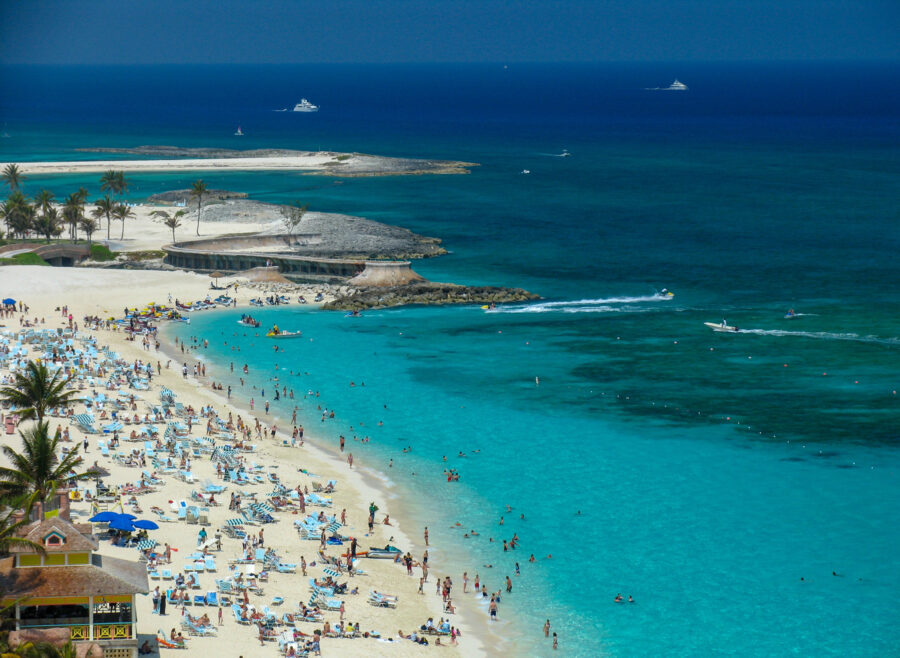 Crowds of beachgoers relax and play on the blue sands of Cable Beach, Nassau, Bahamas, under a clear sky.