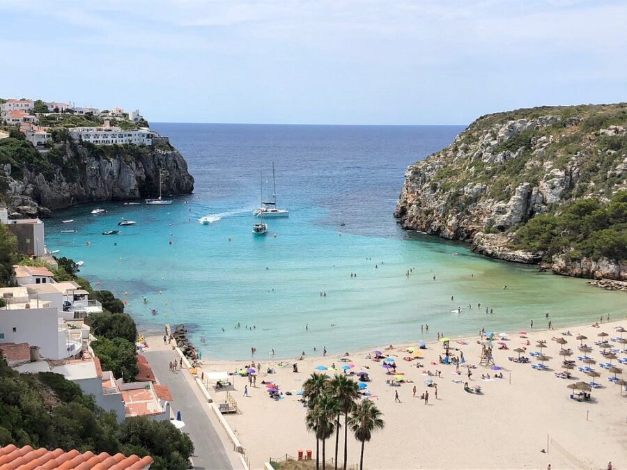 Cala en Porter, Menorca, features a lively beach scene with numerous people and boats dotting the shoreline.