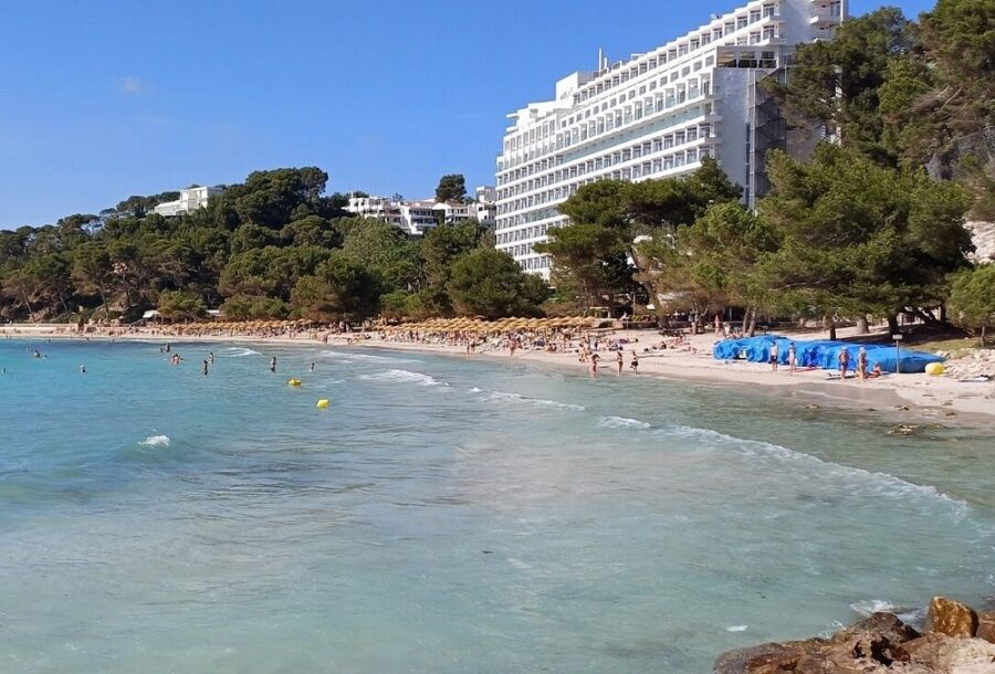 Cala Galdana Beach in Menorca, featuring people swimming with a hotel visible in the background under a clear blue sky.