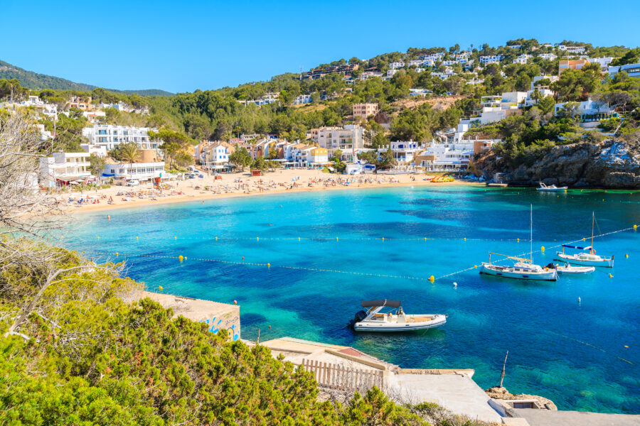 Cala Vadella bay in Ibiza features fishing and sailing boats on clear blue waters, framed by beautiful coastal scenery
