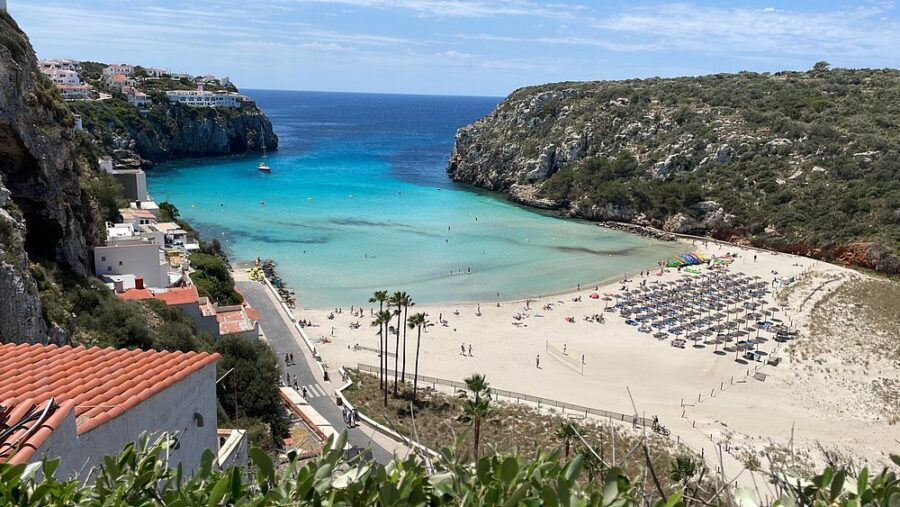 Cala’n Porter beach showcases vibrant blue waters lapping against pristine white sand, framed by a sunny sky.