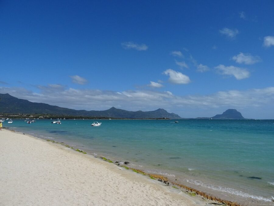 Tropical La Preneuse Beach with white sand, turquoise water, anchored boats, and mountain backdrop.